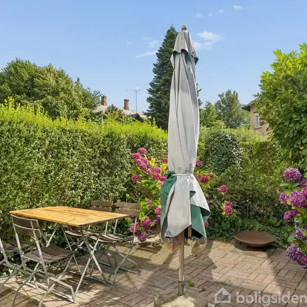 Et udendørs terrasseområde med et bord og stole i træ står på en flisebelagt belægning. En stor parasol er placeret i midten. Omgivet af frodige hække og blomstrende hortensiaer.