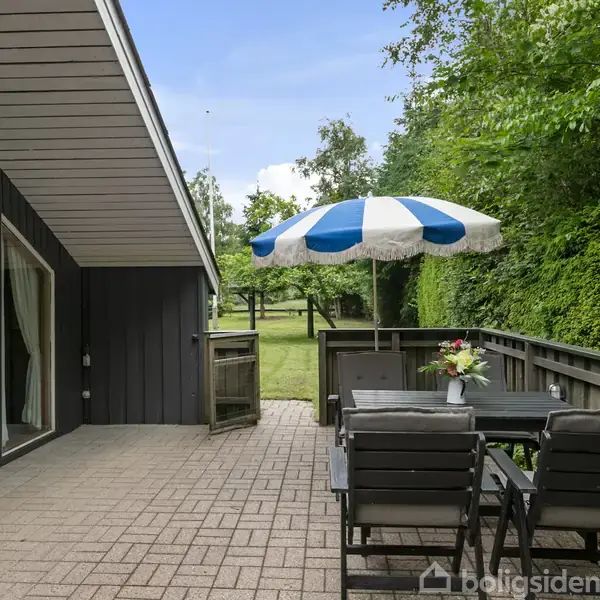 Terrassemøbler står på solrig, flisebelagt terrasse ved mørkt træhus. En blå-hvid parasol giver skygge. Omkranset af tæt hæk og træer, med udsigt til græsplæne i baggrunden.