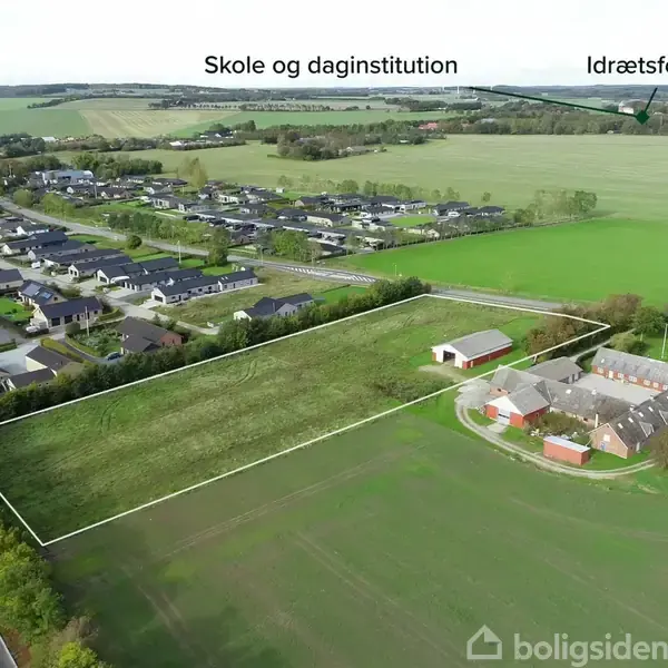 Et stort, rektangulært græsareal ligger ved siden af en mindre bydel med huse. Bygninger ses tæt ved marken. Tekst: "Skole og daginstitution", "Idrætsforening" angiver placeringer i omgivelserne.