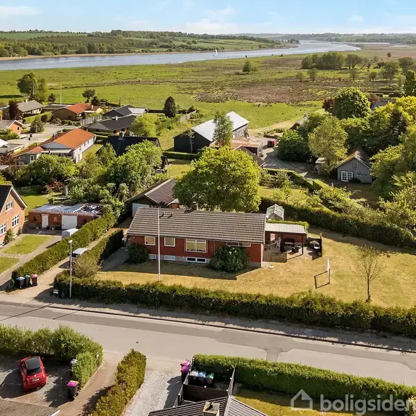Hus med have ligger i en beboelsesområde nær vej, omgivet af andre huse og grønne områder. I baggrunden ses en sø eller flod samt åbne marker. Ingen tekst er synlig.