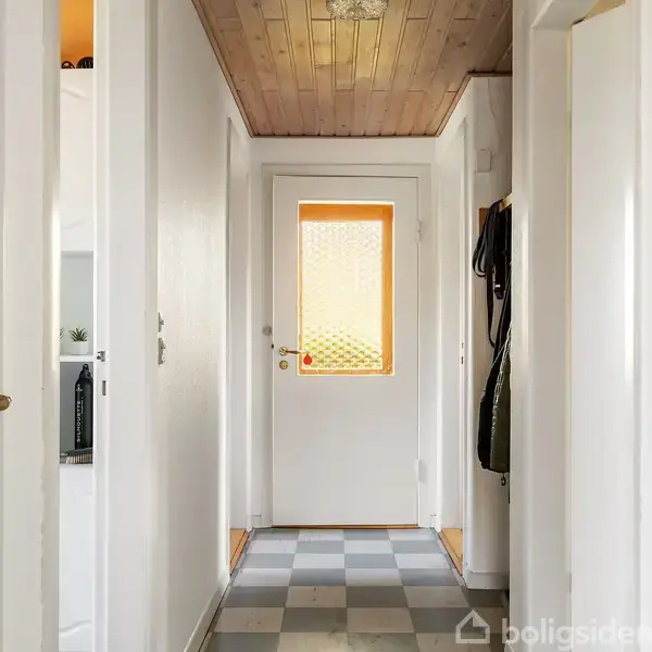 En lang, smal gang med en hvid dør i enden. Døren har et mønstret glasvindue. Væggene er hvide, og gulvet har gråt skakternet fliser. En træloft ses ovenfor.