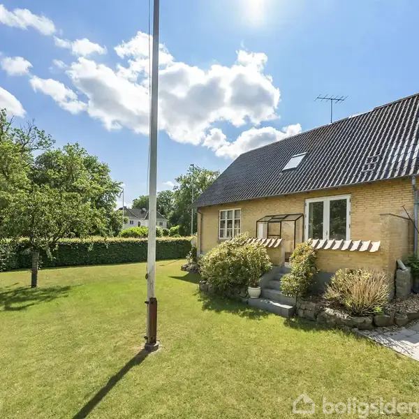 Et gult murstenshus med mørkt tag står i en solrig have med græsplæne, træer og buske. Foran huset ses en flagstang og en flisebelagt terrasse.