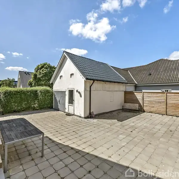 Et flisebelagt gårdhaveområde med et hvidt skur i midten, omgivet af hække og trægavle. Et bord står i forgrunden under en klar blå himmel med få skyer.