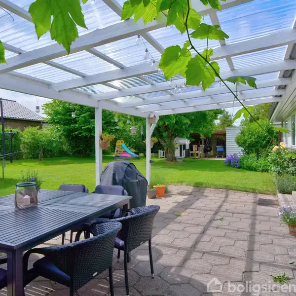 En overdækket terrasse med sorte havemøbler står ved siden af et hus med store vinduer. I baggrunden ses en trampolin og grøn have med legesæt.
