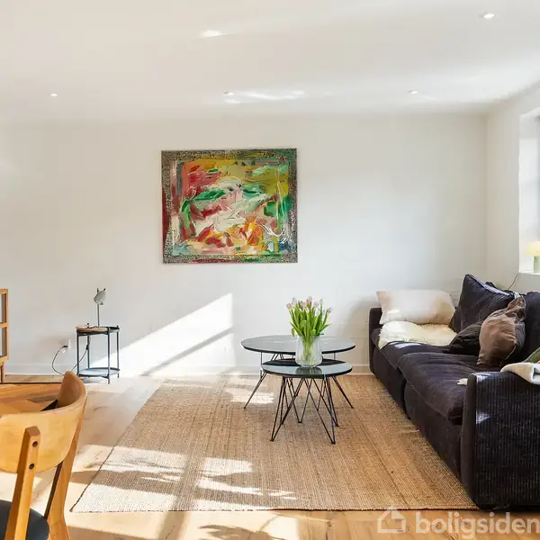 Stue med mørk sofa, dekoreret med puder, står ved vindue. Et rundt sofabord med blomster i vase foran. Abstrakt maleri hænger på lys væg. Fjernsyn på skænk.