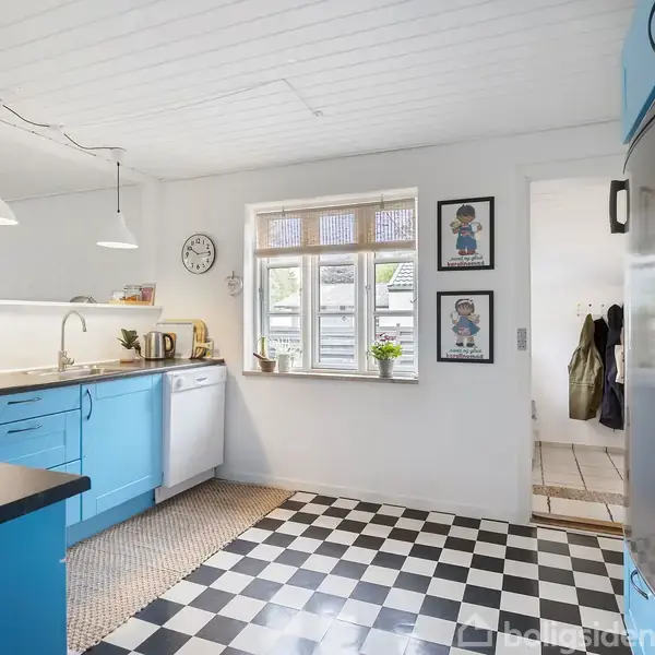 Køkken med lyseblå skabe og checkeret sort-hvidt gulv. Vasen med plante står på bordet. Vægdekorationer hænger ved vinduet; to billeder og en ur.