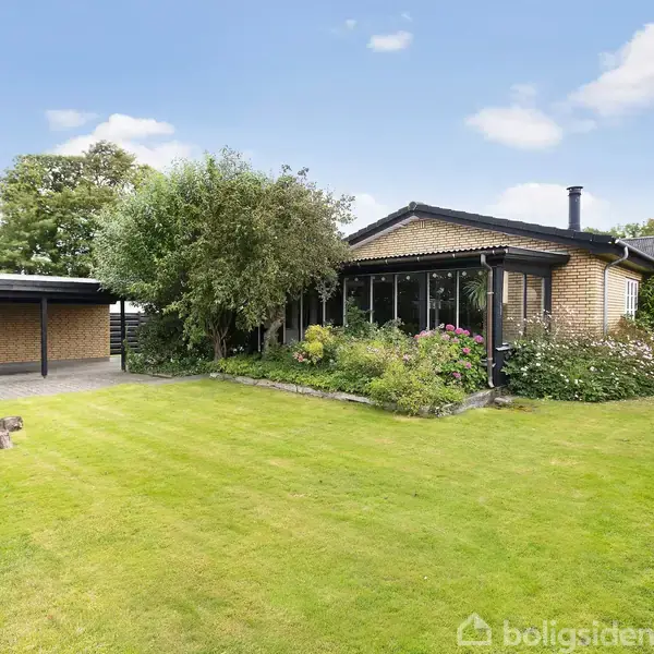 Et én-etagers hus med brune mursten står i en velplejet have med græsplæne og blomsterbede. Til venstre ses en carport og en bålplads med træstubbe som siddepladser.