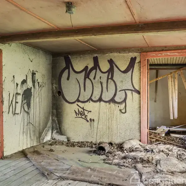 Et forladt rum med graffiti på væggene og skrald på gulvet. Indgangen til et andet rum viser nedbrudte strukturer. Vægge er dekoreret med ordene "KE" og "Net".