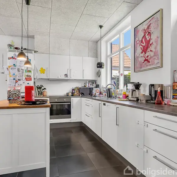 Et moderne køkken med hvide skabe og sorte fliser, hvor køkkenredskaber og apparater er placeret på bordpladerne. Kunst på væggen til højre og hængende lamper over borde.