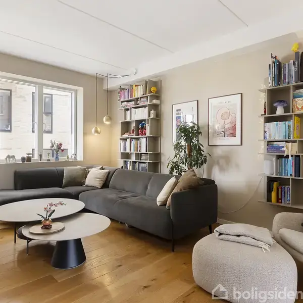 Stue med grå sofa og moderne lænestol ved siden af runde borde. Væggene har malerier og reoler med bøger og planter. Store vinduer slipper lys ind på trægulvet.