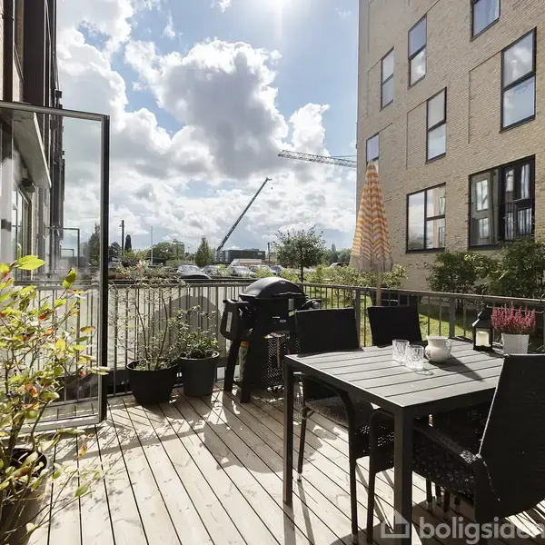 En træterrasse med et udendørs spisebord og stole står ved siden af en grill. Terrassen er omkranset af bygninger med vinduer. Der er blomsterkrukker og bevoksning.