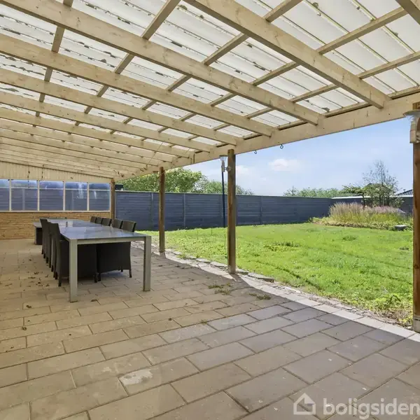 En overdækket terrasse med et langbord og stole på fliser. I haven ved siden af terrassen er der en græsplæne omgivet af et højt hegn under en lysende blå himmel.