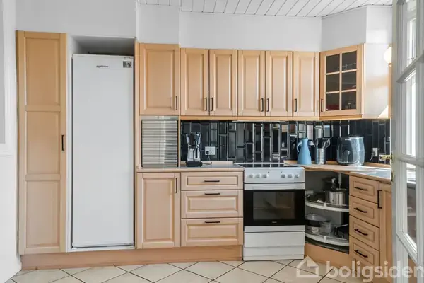 Et køkken med lyst træskab, sort flisebagvæg, en hvid ovn og køleskab. Flere køkkenapparater er placeret på bordpladen. Gulvet har lyse klinker.