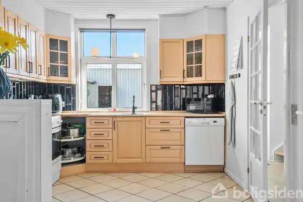 Køkkenet har træskabe, opvaskemaskine, ovn og flisegulv. Det er lyst med et stort vindue og sorte fliser som backsplash. Der hænger en enkel lampe i loftet.