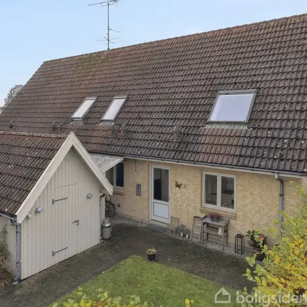 Et hus med skråt tag og flere vinduer står i en have. Der er en havedør og efeu dækker noget af facaden. Omkring ses buske og et lille græsareal.