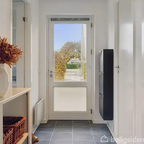 En hvid dør fører til en have med frodige planter. Indendørs står en hvid hylde med en vase fyldt med tørrede blomster. Flisegulvet er i mørke toner.