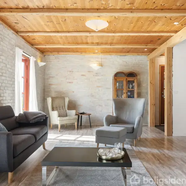 Stue med sofa og lænestol, afslappet indretning. Sofaen har puder, og der står en trækiste ved siden af. Brune murstensvægge og træloft skaber en hyggelig atmosfære.