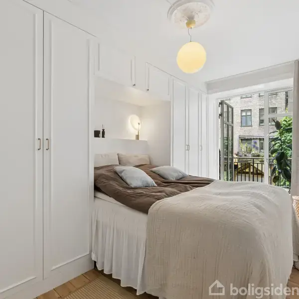 Et soveværelse med en dobbeltseng er omkranset af hvide garderobeskabe. En rund lampe hænger fra loftet. Store vinduer åbner ud til en altan med planter.