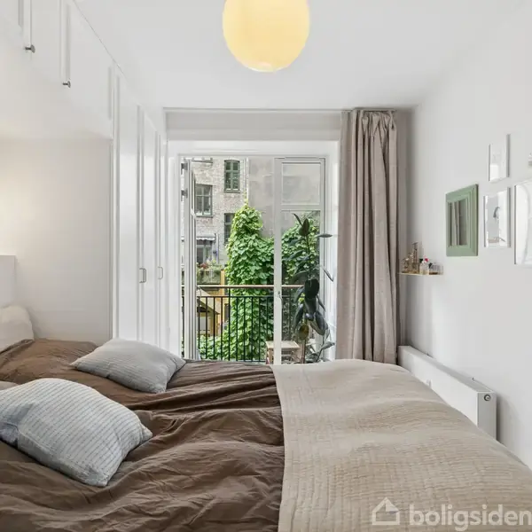 Soveværelse med dobbeltseng dækket med brune og beige sengetæpper. En stor glasdør åbner til en lille balkon med udsigt til grønne planter. Væggene er dekoreret med kunst.