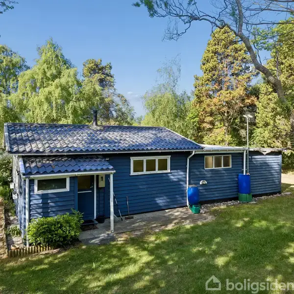 Et blåt træhus står stille i en grøn have omgivet af træer. To regnvandstønder er placeret ved huset, og området ser fredfyldt og solbeskinnet ud.