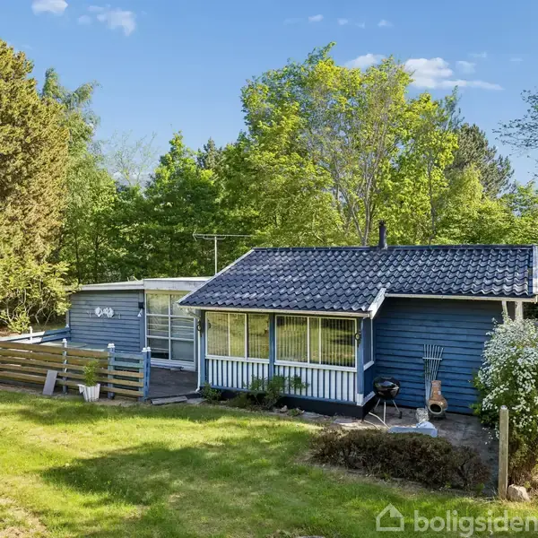 Et blåt træhus med terrasse står i en grøn, skovklædt have. Huset har mørkt tag og store vinduer. Der er en grill foran og høje træer i baggrunden.