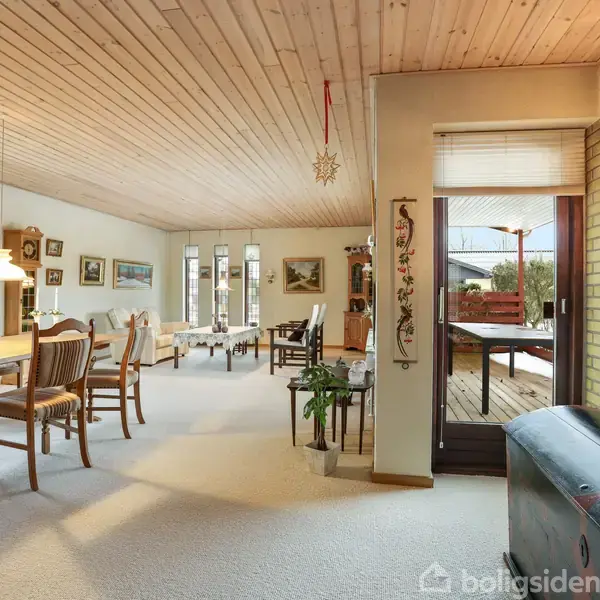 Stue med træloft, spisebord og stole til venstre, sofagruppe i baggrunden. En glasdør fører til en terrasse med havemøbler. Væggene er dekoreret med malerier.