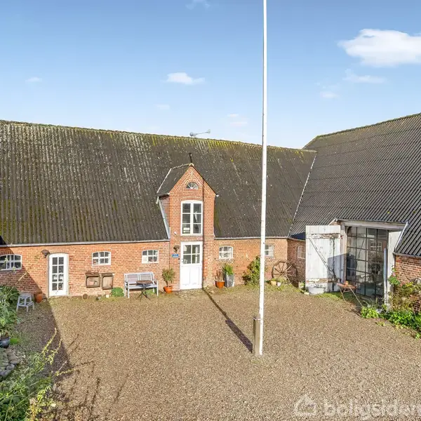 Gårdsplads med grusbelægning omkranset af røde murstenslænger med sorte tagsten; midterbygning har en kvist med dør og vinduer; flagstang centralt placeret; omgivet af grønne planter.