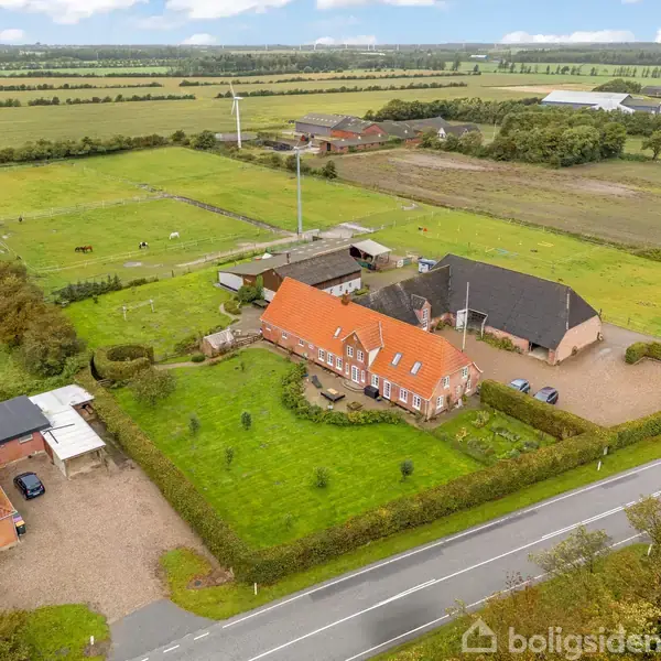 Gårdsetablissement med rødt tegltag bygning og udhus. Omkranset af velplejede græsplæner og hegn i et landligt landskab med heste på markerne i baggrunden. Vejen løber foran.