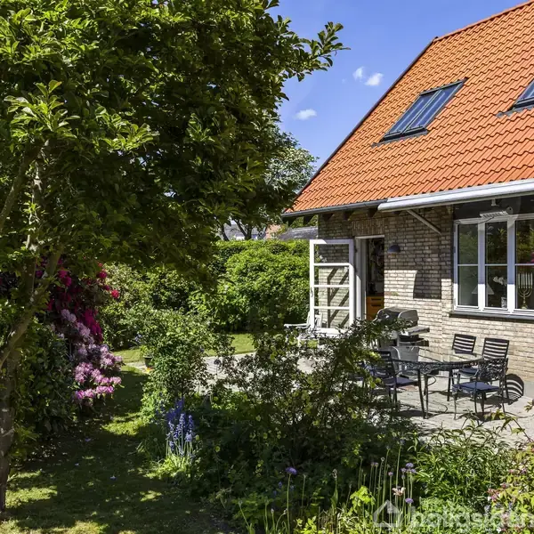 Et hus med rødt tegltag har en terrasse med bord og stole. Haven er frodig med grønne buske og farverige blomster, og solen skinner fra en blå himmel.