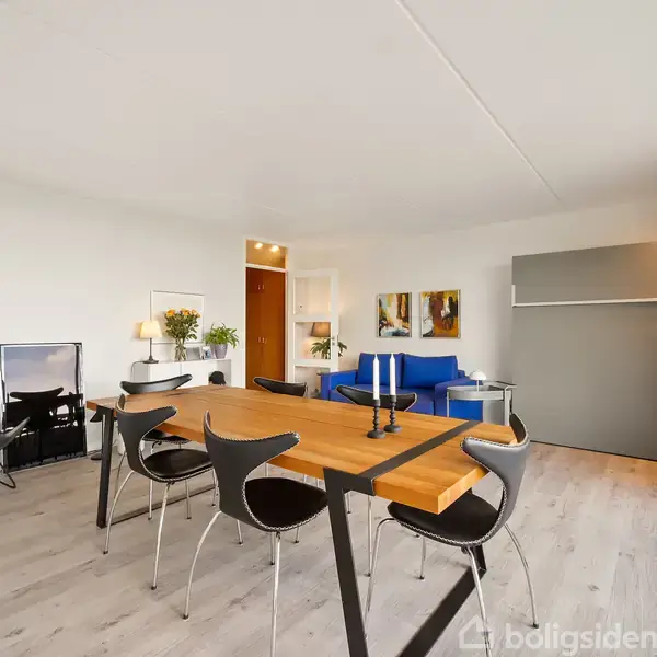 Langt spisebord med sorte stole står i en lys og moderne stue. En blå sofa er placeret ved væggen, dekoreret med malerier. Store vinduer giver udsigt og lysindfald.