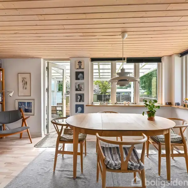 Rundt spisebord omgivet af stole, står på tæppe i lys spisestue med trægulv og bogreol. Store vinduer giver udsigt til have og terrassedør leder udenfor. Lampe hænger over bordet.