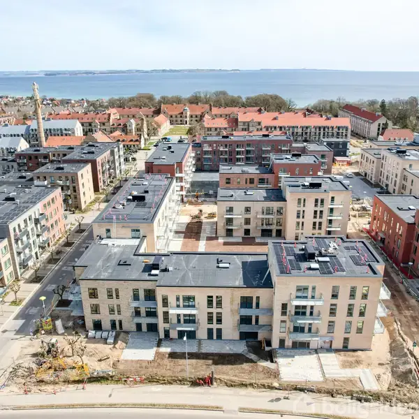 Flere moderne boligbyggerier med flade tage ligger tæt placeret. Bygningerne omgives af veje og er under opførelse. I baggrunden ses træer og en kystlinje med vand.