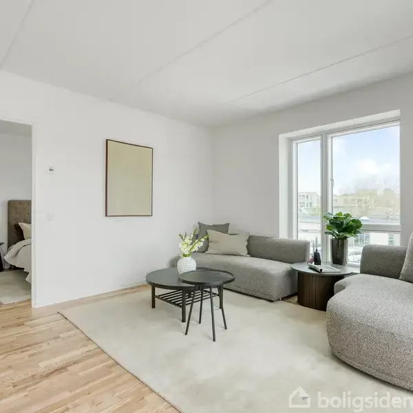 Stue med en grå sofa og et rundt sofabord placeret på et lyst tæppe. Store vinduer slipper naturligt lys ind. I baggrunden ses en åben dør til et soveværelse.