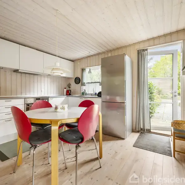 Et køkken med et rundt bord og fire stole i midten. Til venstre er der et hvidt køkkenskab, og til højre en dør mod en terrasse med grønt udenfor.