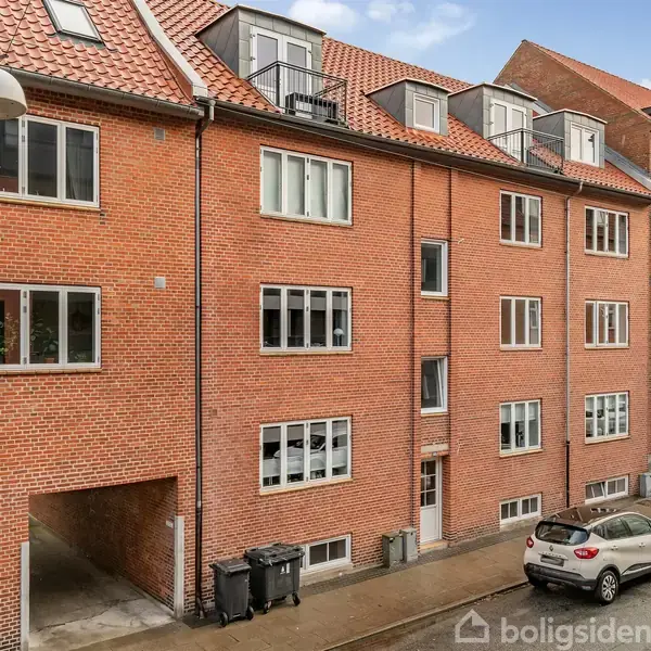 En række rødstensbygninger med skråt tag står stille langs en gade. Biler er parkeret på vejen, og skraldespande står ved en portåbning i bygningen.