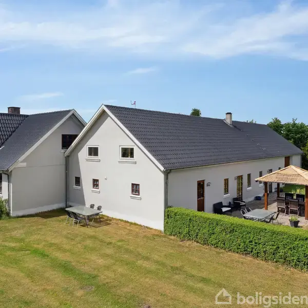 To sammenkoblede hvide bygninger med sorte tage. Foran den højre bygning står et bord og stole ved en terrasse med en pavillon. Græsplænen omgiver husene under en blå himmel.