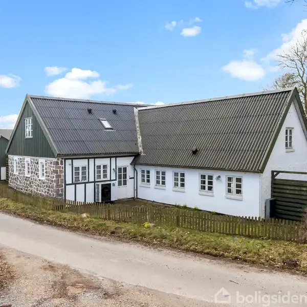 En hvid, traditionel landejendom med sort tag ligger langs en smal landevej, omgivet af marker og træer under en blå himmel med få skyer.