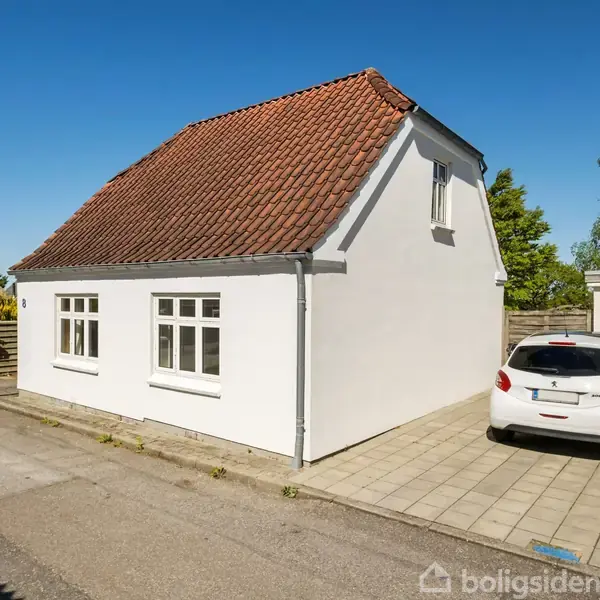 Hvidt hus med rødt tag står stille ved siden af en asfalteret vej. En parkeret hvid bil er placeret på flisebelagt indkørsel med grønne buske og træer i baggrunden.