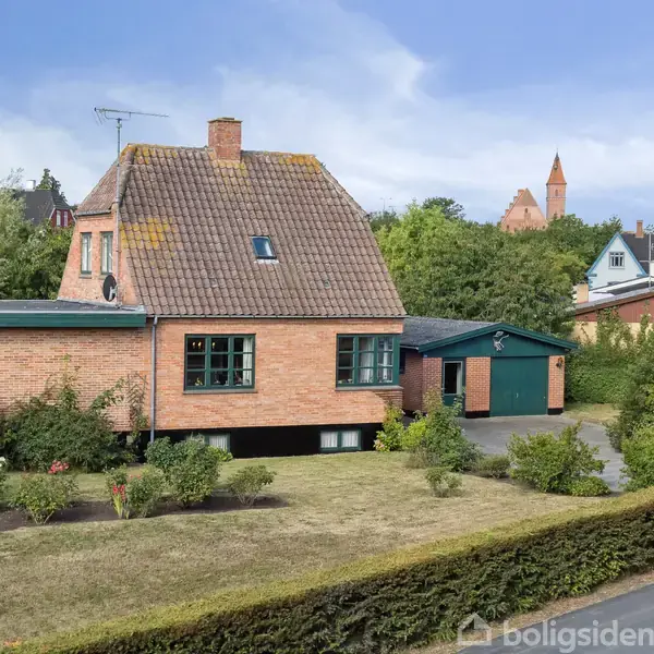 En murstensvilla står i en velplejet have med en indkørsel. Omkring huset er der velholdt græsplæne og buske. I baggrunden ses flere huse og en kirketårn.