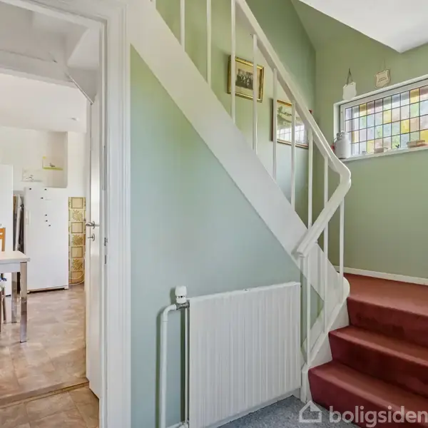 Trappe med rød tæppebeklædning fører opad i en gang med grønne vægge. Til venstre ses et køkken med spisebord og stole. Farvet glasvindue over trappen.