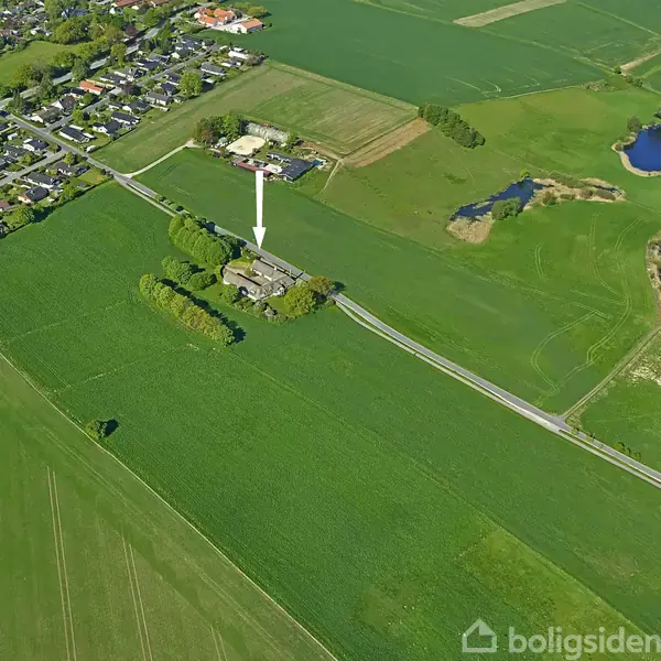 En bygning er peget ud med en pil i et fugleperspektivsbillede. Den ligger ved en vej omgivet af store landbrugsmarker, med en landsby i nærheden.