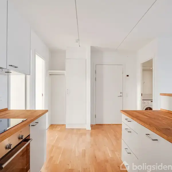 Køkken med hvide skabe og træbordplader, kogeplade og ovn. Rummets lyse hårdttræsgulv strækker sig mod en åben dør og en indgang i et moderne interiør.