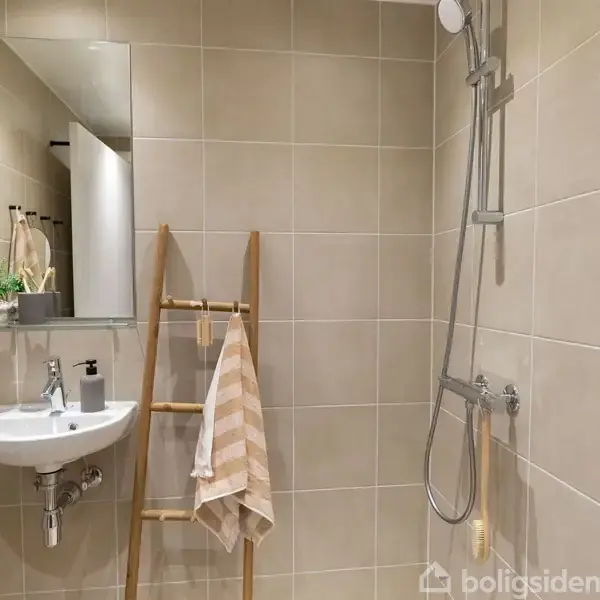 Håndvask med spejl reflekterer badeværelset, dekoreret med lys beige fliser. En stige fungerer som håndklædestang med stribede håndklæder ved siden af en bruser. Planter pynter ved spejlet.