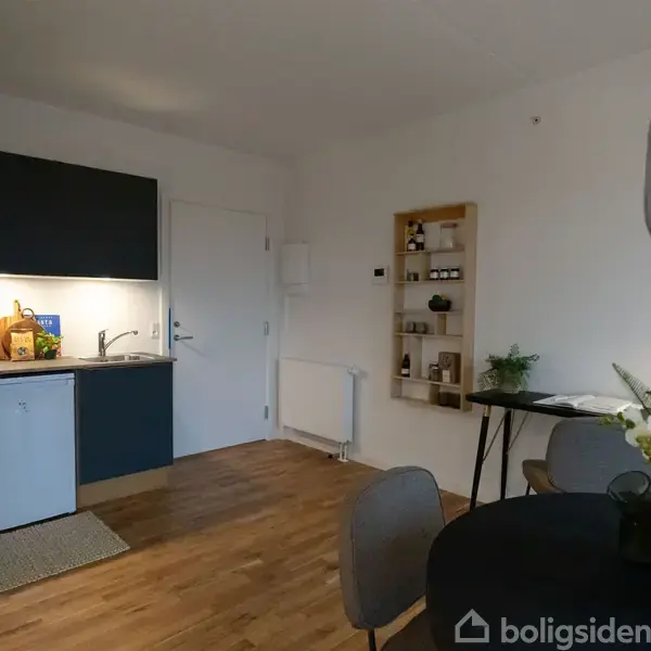 Et moderne køkken med mørke skabe, en vask og komfur til venstre. Rundt spisebord med blomster står til højre. Hylde med dekorationer på væggen. Trægulv.