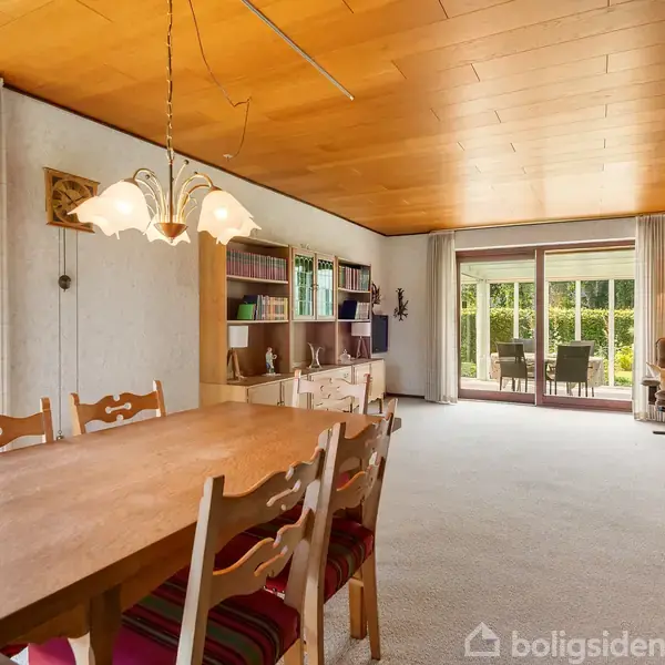 Spisebord med stole, oplyst af lysekrone, i en stue med trægulv. Reoler med bøger står ved siden af. Store vinduer åbner mod udendørs terrasse med yderligere møbler.