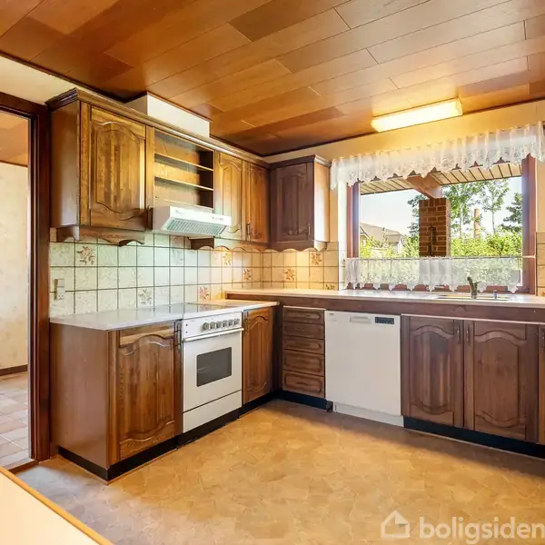 Køkken med brune træskabe, keramisk komfur og opvaskemaskine. Vindue med blondegardin giver udsigt til have. Flisevægge og lyst trægulv skaber varm atmosfære. Åbning mod entréområde.