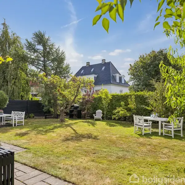 En grøn have med hvide havemøbler placeret på græsset. Træer og buske omringer området. I baggrunden ses et stort, hvidt hus med sort tag under en klar blå himmel.