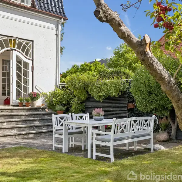 Hvidt havesæt står på flisebelagt terrasse foran et hus med buede dobbeltdøre. Trappen er omgivet af grønne planter og træer under en klar blå himmel.