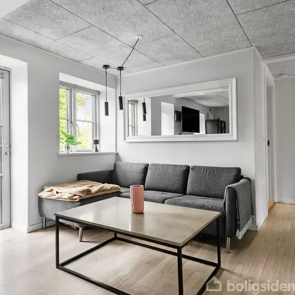 En grå sofa står i stuen med et rektangulært sofabord foran. Et rødt krus står på bordet. Store vinduer giver udsigt til en have, og et stort spejl hænger på væggen.