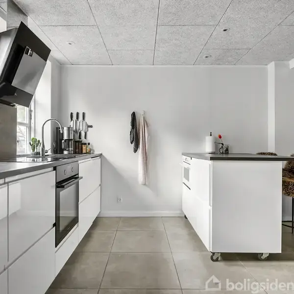 Et moderne køkken med hvide skabe og en central ø. Knive står oprejst, viskestykker hænger på væggen, og stole er placeret ved øen. Lys strømmer ind gennem et vindue.